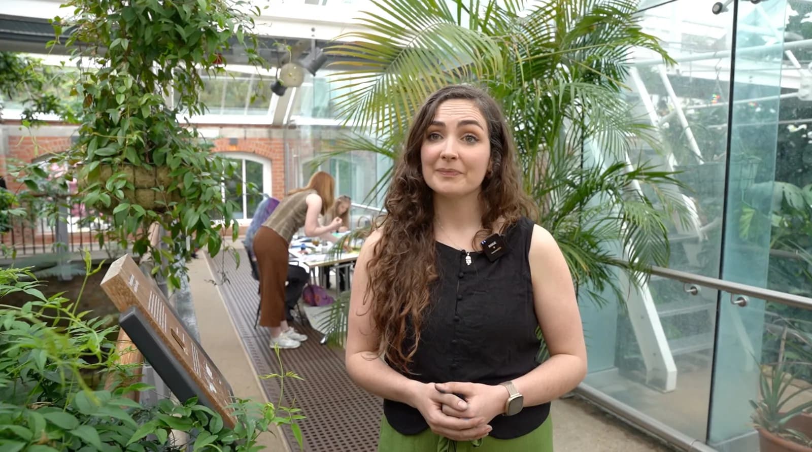 A woman with long, curly brown hair and a black sleeveless top stands in a greenhouse, with lush green plants surrounding her and other people in the background.